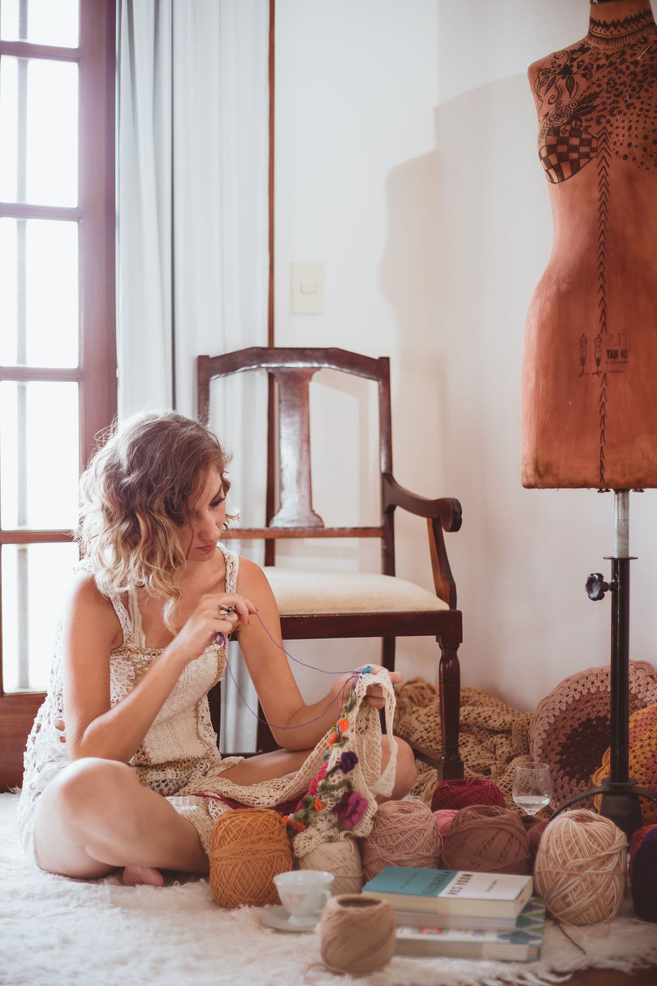 Helena crochetando sentada no chão do ateliê, ao lado do manequim